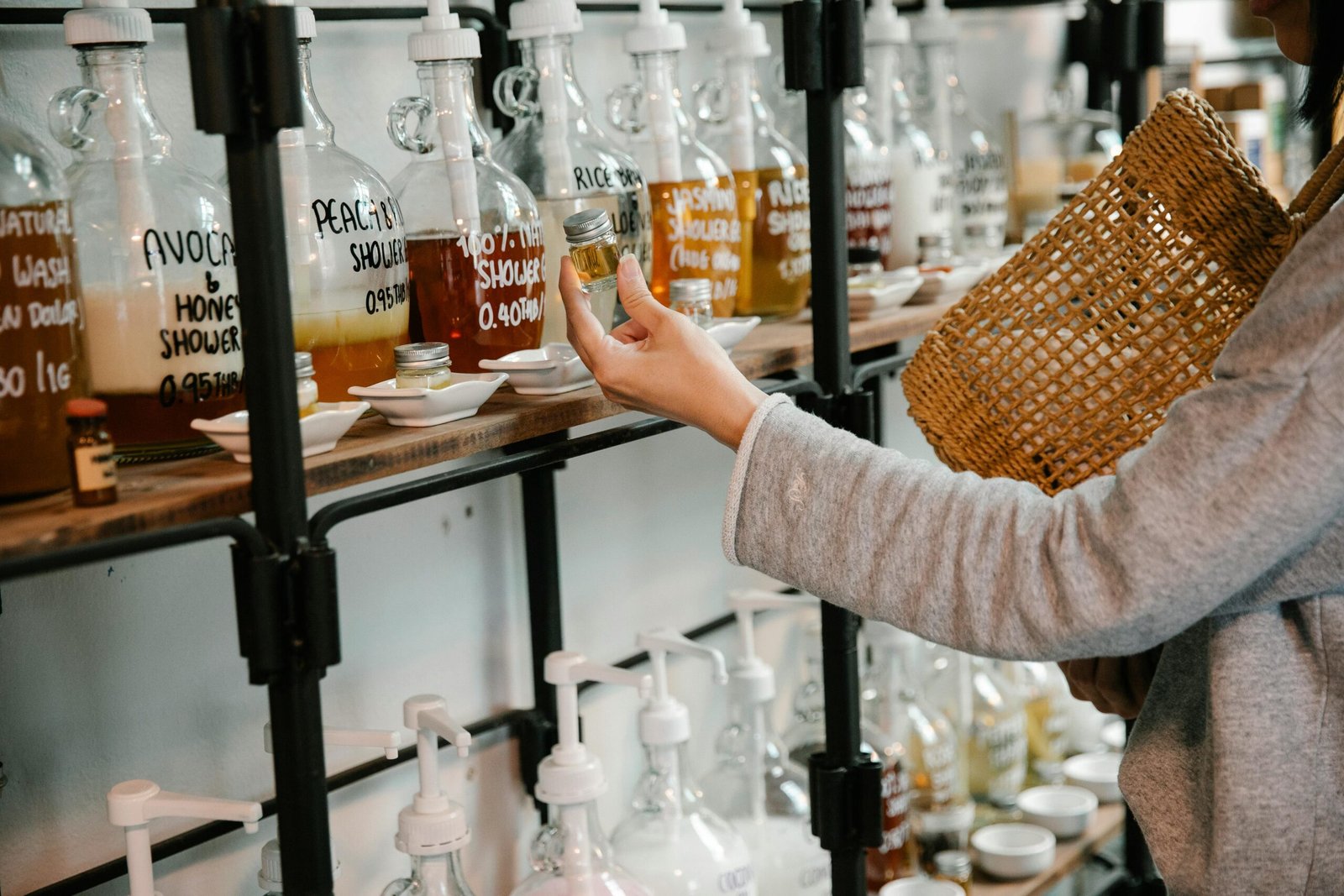 Crop customer examining glass jar with shampoo in market while standing near big dispensers placed on wooden shelves